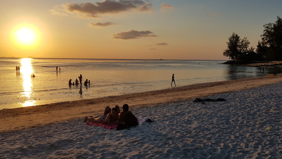 Sunset at the beach, some people are in the shallow water, a couple is laying on a blanket in the sand and watch the sun go down.