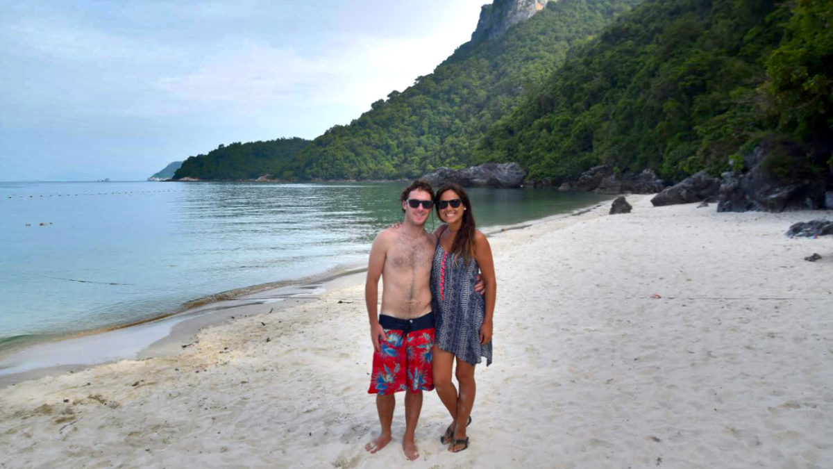 Man and woman standing at the beach smiling at the camera, sun glasses on their eyes, the ocean and a green jungle mountain in the background.
