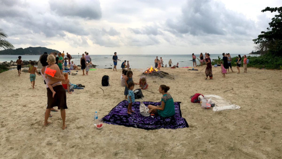people at the beach, in the middle there is a bonfire lighting up, clouds hanging in the sky.