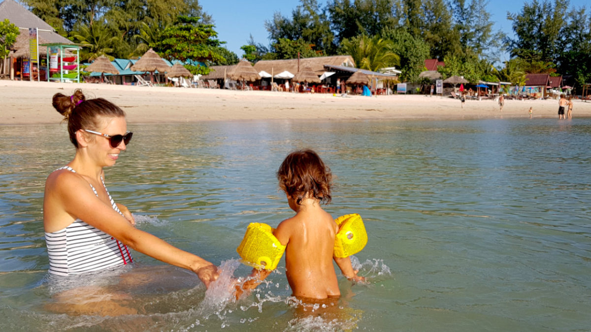 woman with child plays in shallow water at the beach. In the background you can see the sandy part of the beach with little shops and restaurants.