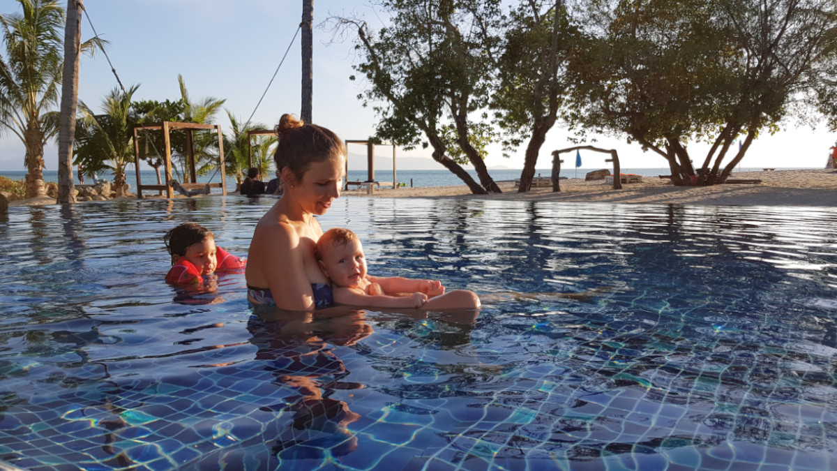 woman swimming with two babies in a swimming pool, the ocean in the background