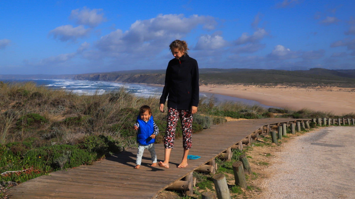 A woman and little child are walking on a pathway, the ocean and beach in the background.