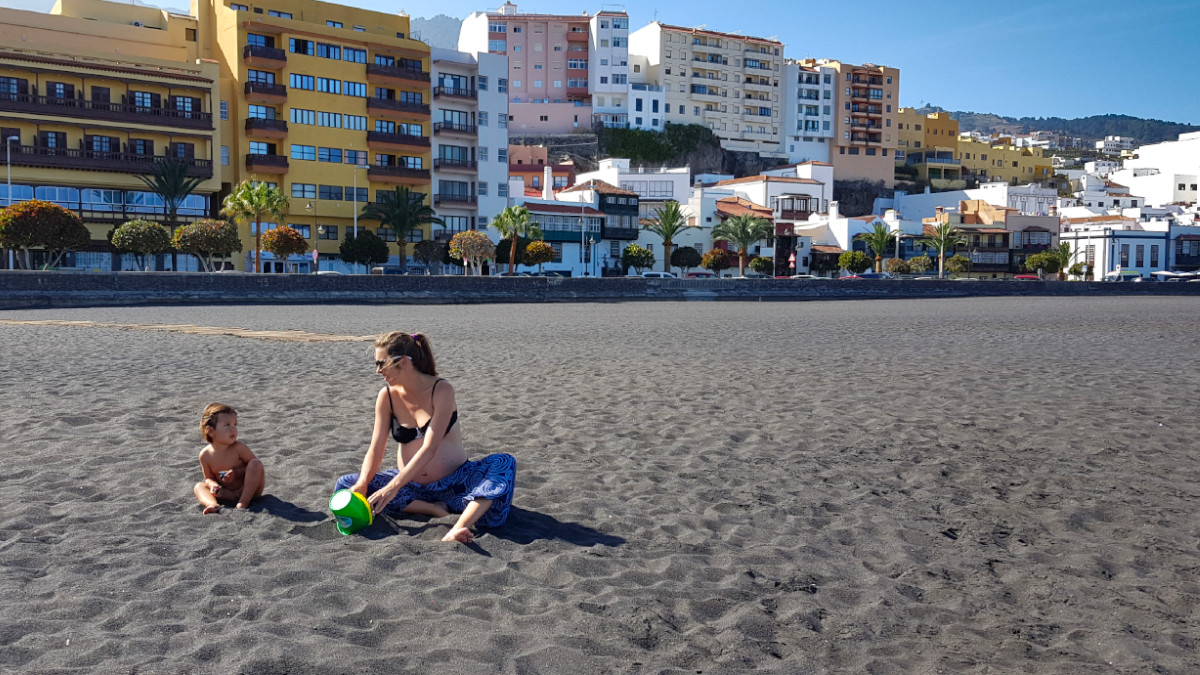 A child and her mother are sitting in the sand of a beach in front of a row of houses.