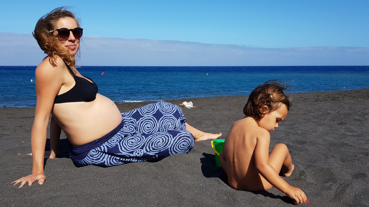 A pregnant woman is sitting in the sand with her son, black sand strand, in the background is the ocean.